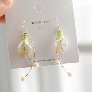 May include: A pair of gold earrings with white flower charms and pearl accents. The earrings are displayed on a white card with the text "THANK YOU" and "Fashion Accessories".