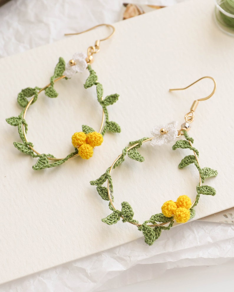 May include: A pair of gold hoop earrings with crocheted green leaves and white and yellow flowers.