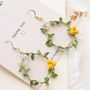 May include: A pair of gold hoop earrings with crocheted green leaves and white and yellow flowers. The earrings are on a white background with the text "Thank You" written on it.