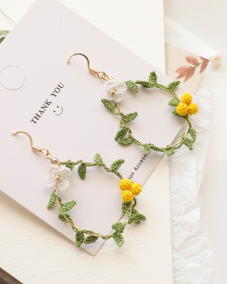 May include: A pair of gold hoop earrings with crocheted green leaves and white and yellow flowers. The earrings are on a white background with the text "Thank You" written on it.