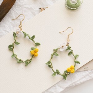 May include: A pair of gold hoop earrings with crocheted floral designs. The earrings feature white flowers and green leaves, with yellow flower buds.