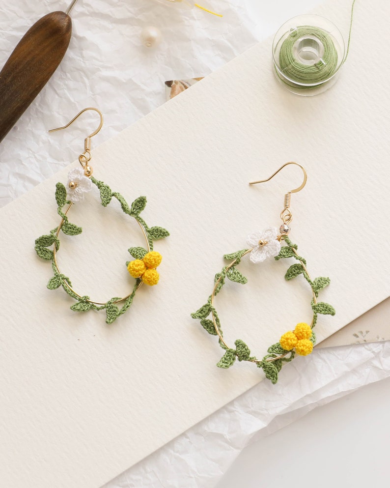 May include: A pair of gold hoop earrings with crocheted floral designs. The earrings feature white flowers and green leaves, with yellow flower buds.