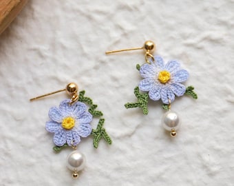 May include: A pair of delicate earrings featuring crocheted lavender flowers with yellow centers and green leaves, each dangling from a gold post and a pearl.