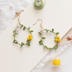 May include: A pair of gold hoop earrings with crocheted floral designs. The earrings feature green leaves and white and yellow flowers.