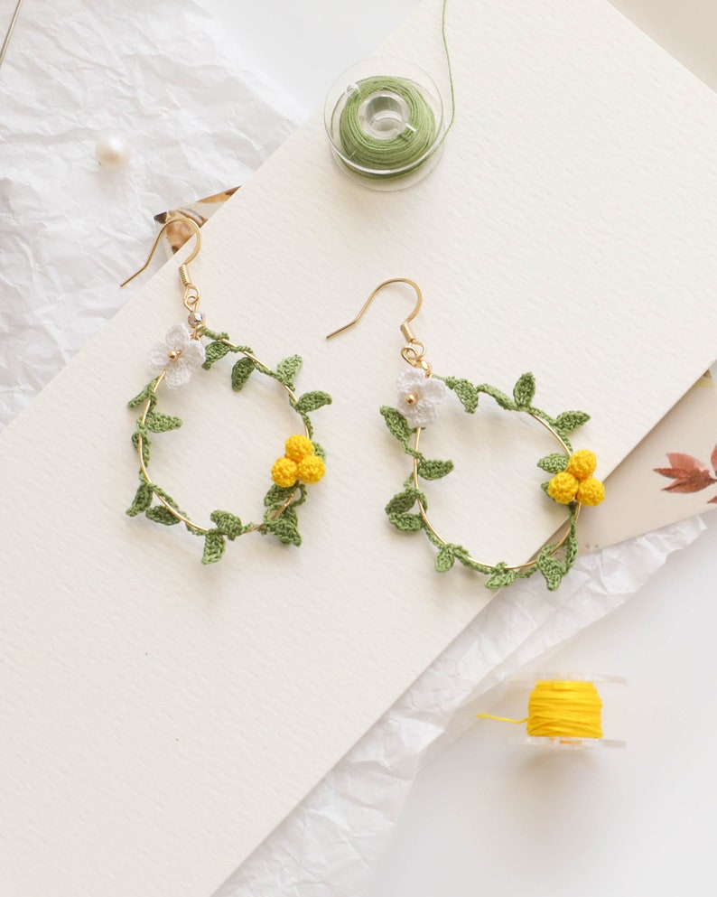 May include: A pair of gold hoop earrings with crocheted floral designs. The earrings feature green leaves and white and yellow flowers.