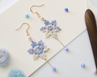 May include: A pair of crocheted snowflake earrings with blue and white accents. The earrings are hanging from gold hooks and have blue beads dangling from the bottom.