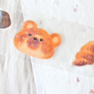 May include: Close-up of a sheet of stickers featuring a bear and a shell design. The bear sticker is orange with star accents and black eyes. The shell sticker is orange and brown. The stickers are on a clear backing.