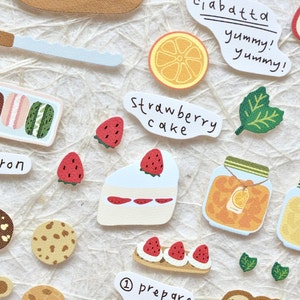 Op de afbeelding: Een verzameling kleurrijke stickers met verschillende voedingsmiddelen, waaronder macarons, koekjes, een aardbeientaart, een pot oranje marmelade en een brood. De stickers bevatten ook tekst zoals "macaron", "aardbeientaart" en "ciabatta lekker! lekker!"