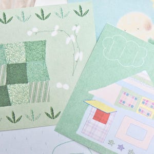 May include: A collection of illustrated cards with a pastel color palette. One card features a house with a blue roof and red door. Another card has a patchwork design in shades of green. The cards have a whimsical, artistic style.