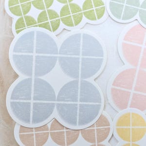 Könnte beinhalten: Eine Sammlung dekorativer Aufkleber in verschiedenen Pastellfarben, darunter Blau, Grün, Rosa und Gelb. Jeder Aufkleber hat ein kreisförmiges Design mit einem Kreuzmuster, das ein gitterartiges Aussehen erzeugt. Die Aufkleber haben eine weiche, strukturierte Optik.