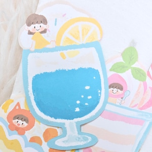 Puede incluir: Recortes de papel coloridos de bebidas y personajes de dibujos animados. Una bebida azul con una rodaja de limón y una niña con un vestido amarillo. Otros recortes incluyen un postre de melocotón y un personaje con un atuendo naranja. El tema general es lindo y caprichoso.