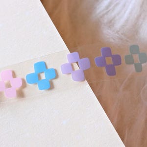 以下が含まれることがあります： パステルカラーの十字架が描かれた透明な和紙テープのロール。十字架はピンク、ブルー、パープル、グリーン、イエローのカラーで構成されています。
