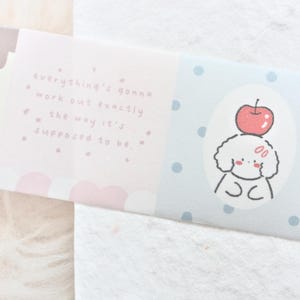 May include: A decorative ribbon with a pastel color scheme. The ribbon features a cartoon poodle with an apple on its head, and the text "everything's gonna work out exactly the way it's supposed to be."