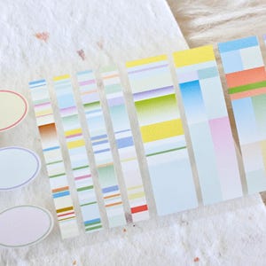 Op de afbeelding: Een set kleurrijk washi tape met geometrische patronen en verlopen in tinten blauw, groen, geel, roze en wit. De tape is gerangschikt in rijen op een wit, getextureerd oppervlak.