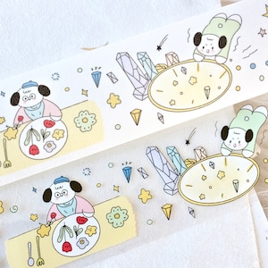 Op de afbeelding: Twee rollen transparante washi tape met kleurrijke illustraties. Op de ene rol staat een hond die een bord met bloemen schildert, terwijl op de andere rol een hond een klok vasthoudt met sterren en kristallen eromheen.