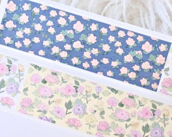May include: Two rolls of washi tape with floral patterns. One roll has a light blue background with pink flowers. The other roll has a light yellow background with pink and purple flowers.
