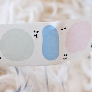 May include: A white washi tape with different colored shapes and faces. The shapes are a light purple cloud, a light green circle, a light blue blob, and a light pink blob. Each shape has a different face drawn on it.