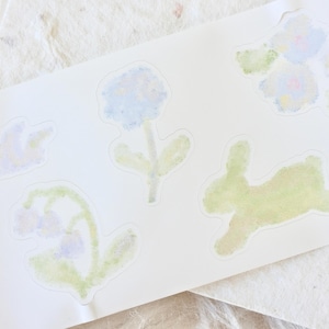 May include: A sheet of watercolour stickers featuring floral and bunny designs. The stickers are in soft pastel colours of blue, green, and pink. The sheet is on a white background.