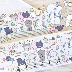 May include: A sheet of white washi tape with a repeating pattern of cute cartoon dogs and cats playing musical instruments. The tape features a variety of colors, including pink, blue, yellow, and black. The tape is decorated with musical notes and stars.