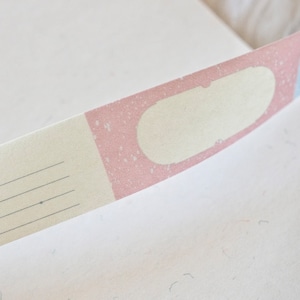 Op de afbeelding: Een witte en roze washi tape met een leeg ovaal label. De tape heeft een subtiel gespikkeld patroon en een dun zwart lijntjesontwerp.