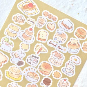 May include: A sheet of stickers featuring cute cartoon illustrations of food and bears. The stickers are on a brown background and include images of bread, coffee, pastries, and other treats. The text "Bread Bear" is included on the sheet.