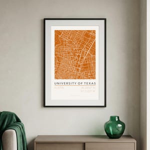 Puede incluir: Impresión enmarcada del mapa de la Universidad de Texas en Austin. El mapa es de color naranja quemado, con las calles y edificios de la ciudad. La impresión está en un marco negro y cuelga de una pared gris claro.