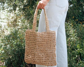 May include: A woven tote bag with a rectangular shape and two handles. The bag is made of a light brown material with a textured, open weave design. The bag is being held in front of a background of green foliage.