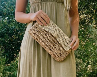 May include: A light brown woven clutch with a flap closure is held in front of a light olive green dress. The dress has a smocked bodice and spaghetti straps. The background is a lush green bush.