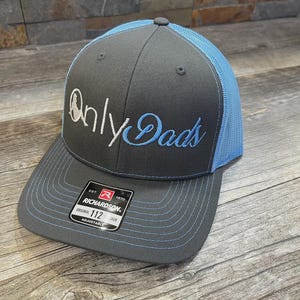 May include: A gray and blue trucker hat with the text "Only Dads" embroidered in white and blue. The hat features a curved brim and a mesh back. The Richardson brand label is visible.
