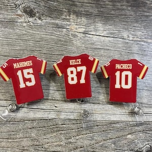 May include: Three red football jersey-shaped badge reels. Each reel features a different player's name and number: Mahomes 15, Kelce 87, and Pacheco 10. The jerseys have gold accents and white numbers. The reels are on a wooden surface.