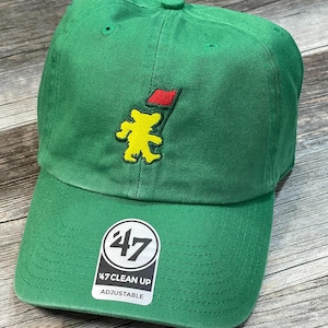 May include: Green adjustable baseball cap with a yellow bear holding a red flag. The cap has a curved brim and a 47 Clean Up Adjustable sticker.