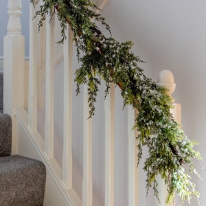 Könnte beinhalten: Ein festlicher Girlande aus grünen Tannenzweigen mit Schneebesatz schmückt ein Treppengeländer. Die Girlande ist diagonal über dem weißen Geländer drapiert und verleiht dem Innenraum eine festliche Note, ideal für die Weihnachtszeit.