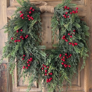 May include: A Christmas wreath featuring evergreen foliage and bright red berries. The wreath is suspended by a rope loop and is set against a wooden door. A traditional festive decoration for the holiday season.
