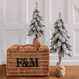 Op de afbeelding: Twee kleine, besneeuwde, kunstkerstbomen met jute bases staan voor een witte paneeldeur. Een bruine rieten mand met de letters "F&M" gedrukt op de voorkant staat op de houten vloer.
