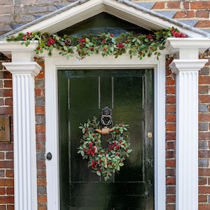 May include: A dark green door adorned with a festive wreath and garland, framed by white pillars and a pediment. The wreath and garland are decorated with red berries and green leaves, set against a brick wall.
