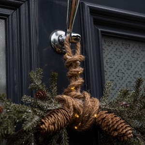 Può includere: Una ghirlanda di corda marrone con rami di pino e pigne, decorata con piccole luci bianche, appesa a una porta nera con un batacchio d'argento.