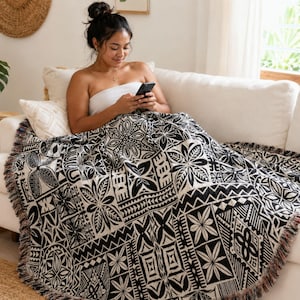 May include: A woven throw blanket featuring a black and white geometric pattern with repeating floral and tribal motifs. The rectangular blanket has a fringed edge and is draped over a white sofa, displaying a high-contrast design throughout.