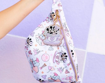 May include: A white fanny pack featuring a repeating pattern of Dumbo, Mickey Mouse, and Minnie Mouse in teacups, castles, and balloons in shades of pink and purple. A gold-toned metal charm of Mickey and Minnie in a teacup hangs from the zipper.