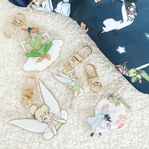 Puede incluir: Llaveros acrílicos con los personajes de Peter Pan y Campanilla. Los llaveros tienen cierres dorados y se muestran con una riñonera azul marino con un tema de Peter Pan. Los llaveros están en varias poses y diseños.