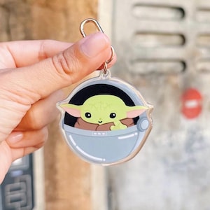 Pu&ograve; includere: Un portachiavi in acrilico trasparente con un'immagine a cartoni animati di Grogu, noto anche come Baby Yoda, in un marsupio azzurro. Il portachiavi ha un anello color oro per attaccarlo a chiavi o borse. Il personaggio ha grandi orecchie verdi e occhi marroni.