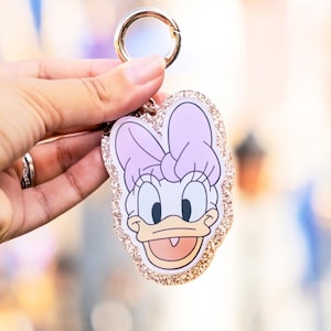 May include: A Daisy Duck keychain with a gold-colored clasp. The keychain features a cartoon image of Daisy Duck with a pink bow, a white face, and a glittery gold border. The keychain is held up by a hand.