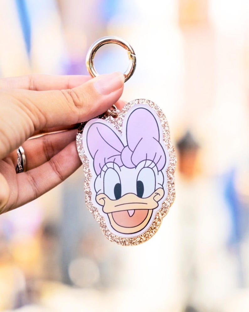 May include: A Daisy Duck keychain with a gold-colored clasp. The keychain features a cartoon image of Daisy Duck with a pink bow, a white face, and a glittery gold border. The keychain is held up by a hand.