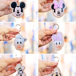 May include: A collection of character keychains featuring Mickey Mouse, Minnie Mouse, Donald Duck, Daisy Duck, and Goofy. Each keychain has a gold glitter border and a gold keyring. The characters are in color with black, white, pink, blue, and yellow.
