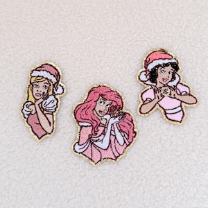 May include: Three embroidered iron-on patches featuring cartoon princesses wearing Santa hats. The princesses are wearing pink dresses and have blonde, pink, and brown hair.