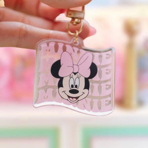 May include: A clear acrylic keychain featuring Minnie Mouse. The keychain has a pink background with the word "MINNIE" repeated. Minnie Mouse is depicted with a pink bow and smiling face. The keychain is attached to a gold-colored metal clasp and a pink strap.