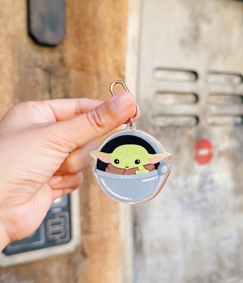 Pu&ograve; includere: Un portachiavi in acrilico trasparente con un'immagine a cartoni animati di Grogu, noto anche come Baby Yoda, in un marsupio azzurro. Il portachiavi ha un anello color oro per attaccarlo a chiavi o borse. Il personaggio ha grandi orecchie verdi e occhi marroni.
