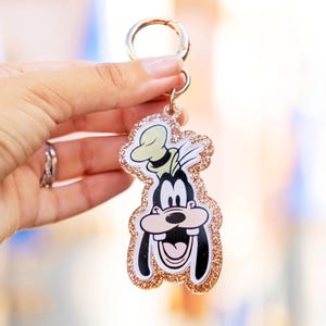 May include: A Goofy-shaped keychain with a gold glitter border and a gold-colored clasp. The cartoon character is smiling with a black and white face, a green hat, and a pink tongue. The keychain is held up against a blurred background.