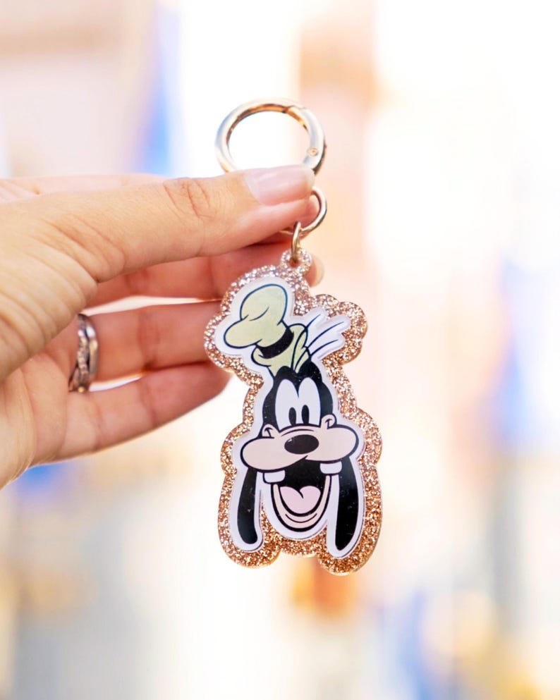 May include: A Goofy-shaped keychain with a gold glitter border and a gold-colored clasp. The cartoon character is smiling with a black and white face, a green hat, and a pink tongue. The keychain is held up against a blurred background.