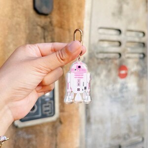 Pu&ograve; includere: Un portachiavi in acrilico trasparente con un design a robot rosa e bianco. Il portachiavi ha un fermaglio in metallo color oro. Il design del robot &egrave; una rappresentazione stilizzata di un popolare personaggio di fantascienza.