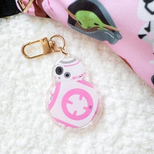 Pu&ograve; includere: Un portachiavi BB-8 in acrilico trasparente con chiusura color oro. Il droide &egrave; bianco con dettagli rosa e un occhio nero. Lo sfondo include un tessuto rosa a tema Star Wars.